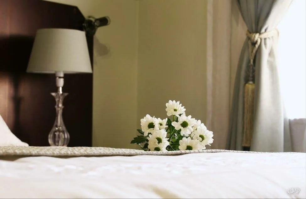 A small bunch of white daisies on a made-up bed, with a bedside lamp and curtain behind
