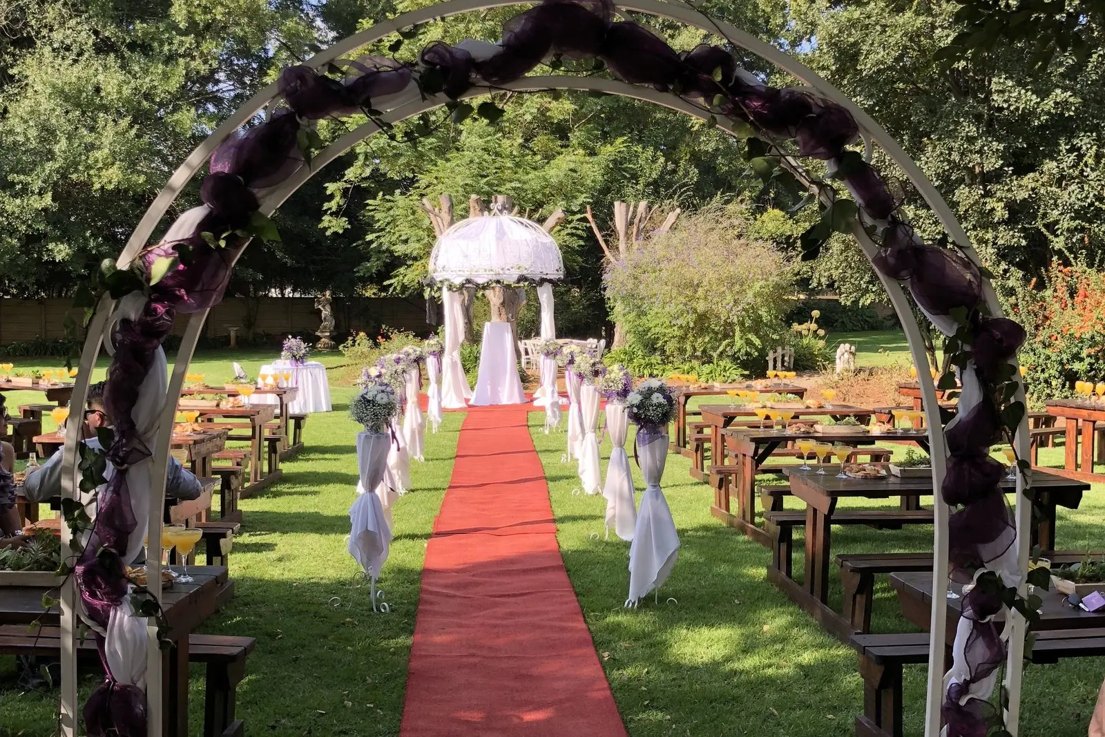 A garden wedding ceremony with a green floral arch, red carpet aisle and covered chairs